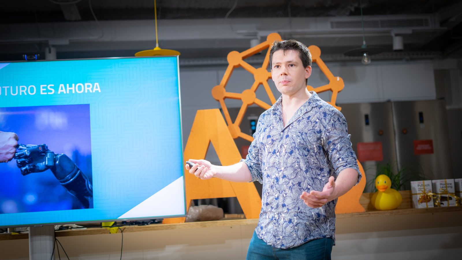 David Robert giving a talk beside a screen showing a robotic hand.
